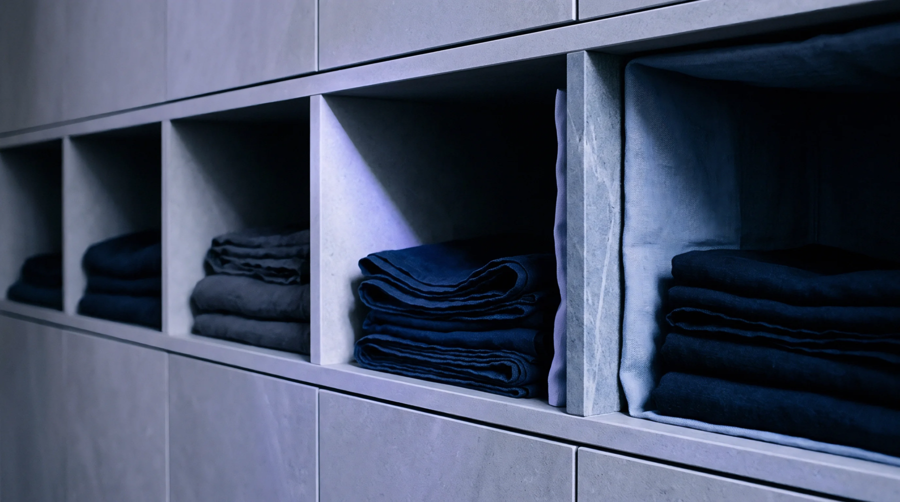Neatly folded stacks of dark blue and grey linen textiles are precisely organized within recessed compartments of a light grey storage system.