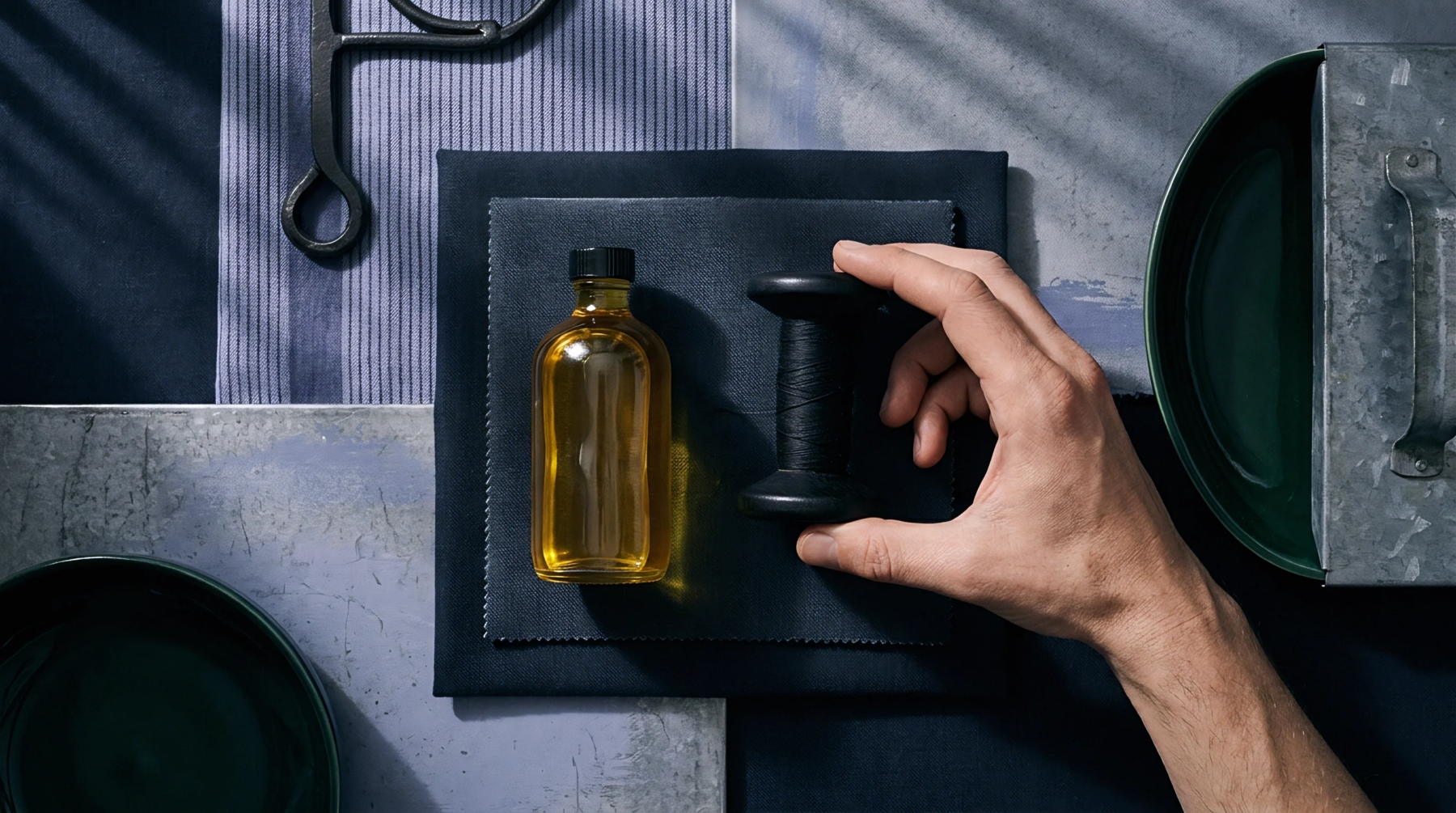 A human hand touches a black wooden spool of thread resting beside a glass bottle of amber oil on dark fabric swatches.
