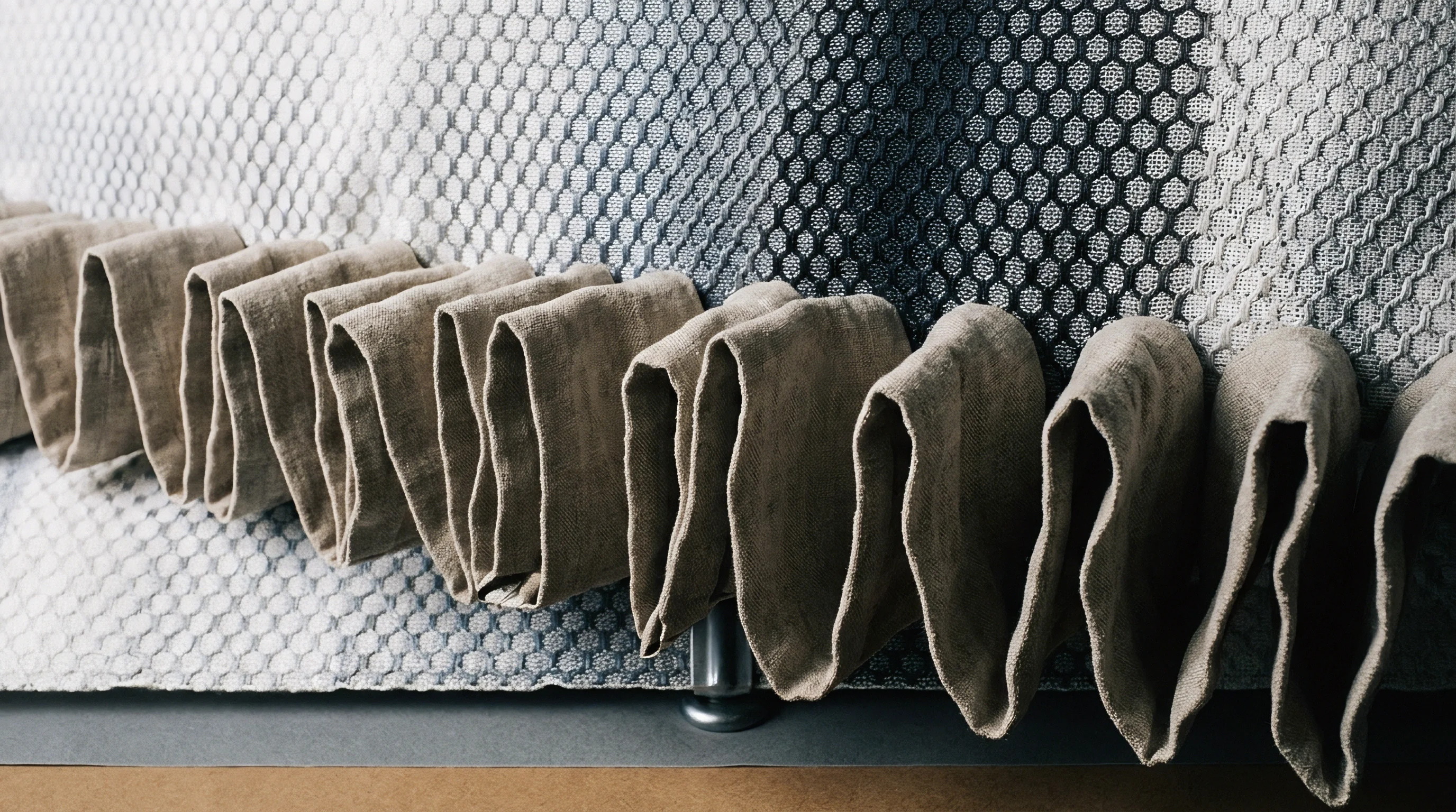 Heavy flax fabric hangs in deep rhythmic folds along a metal support rod beneath textured honeycomb weave panels.