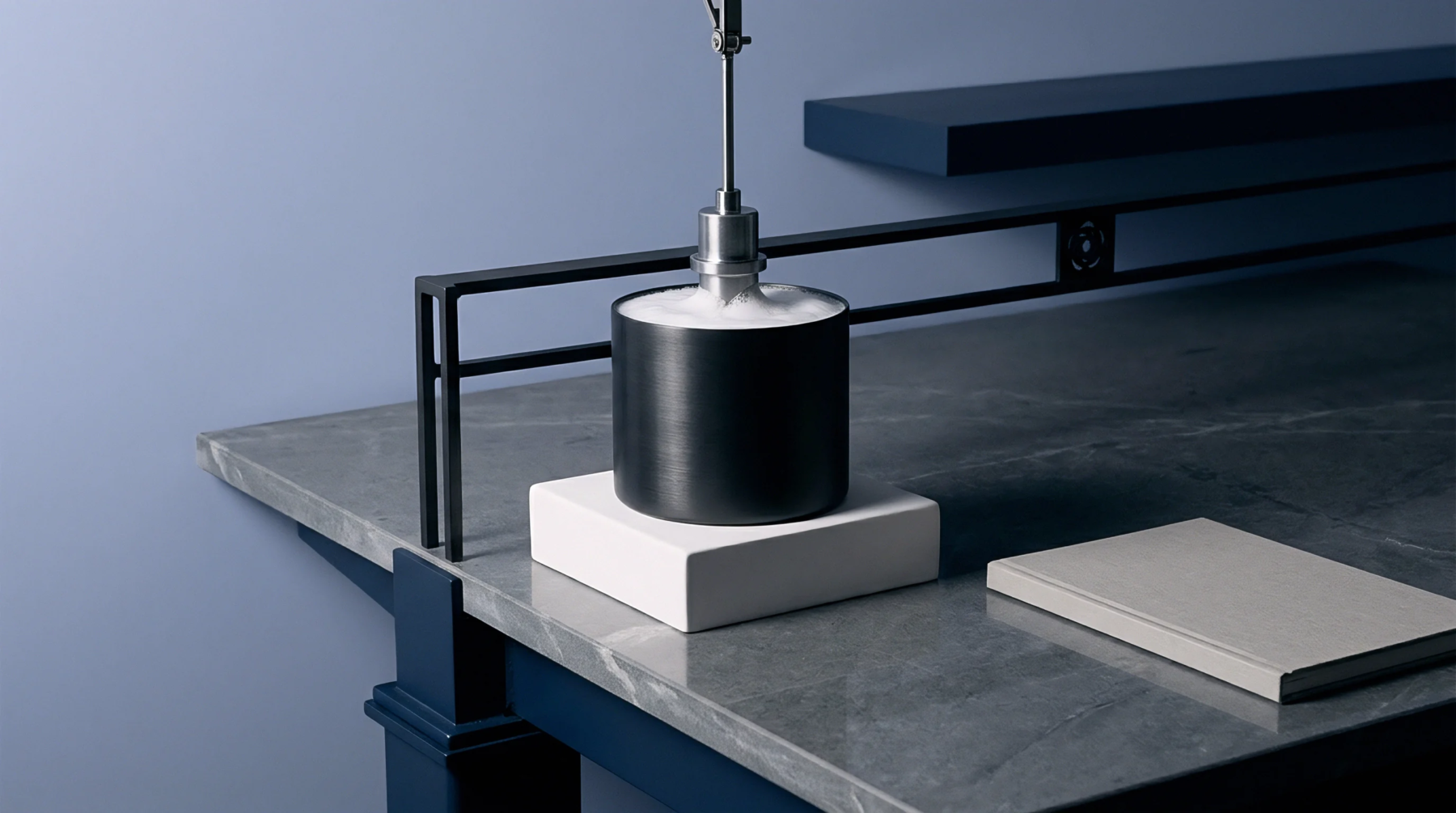 A vertical mechanical spindle agitates soapy liquid inside a cylindrical metal vessel resting on a white stone block upon a workbench.