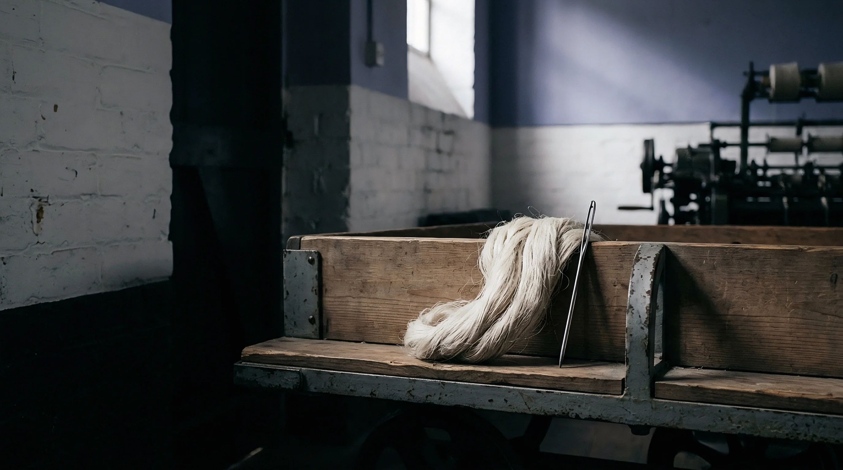Raw flax fibre sits atop a weathered wooden trolley inside a dim textile manufacturing facility with mechanical equipment visible in the background.