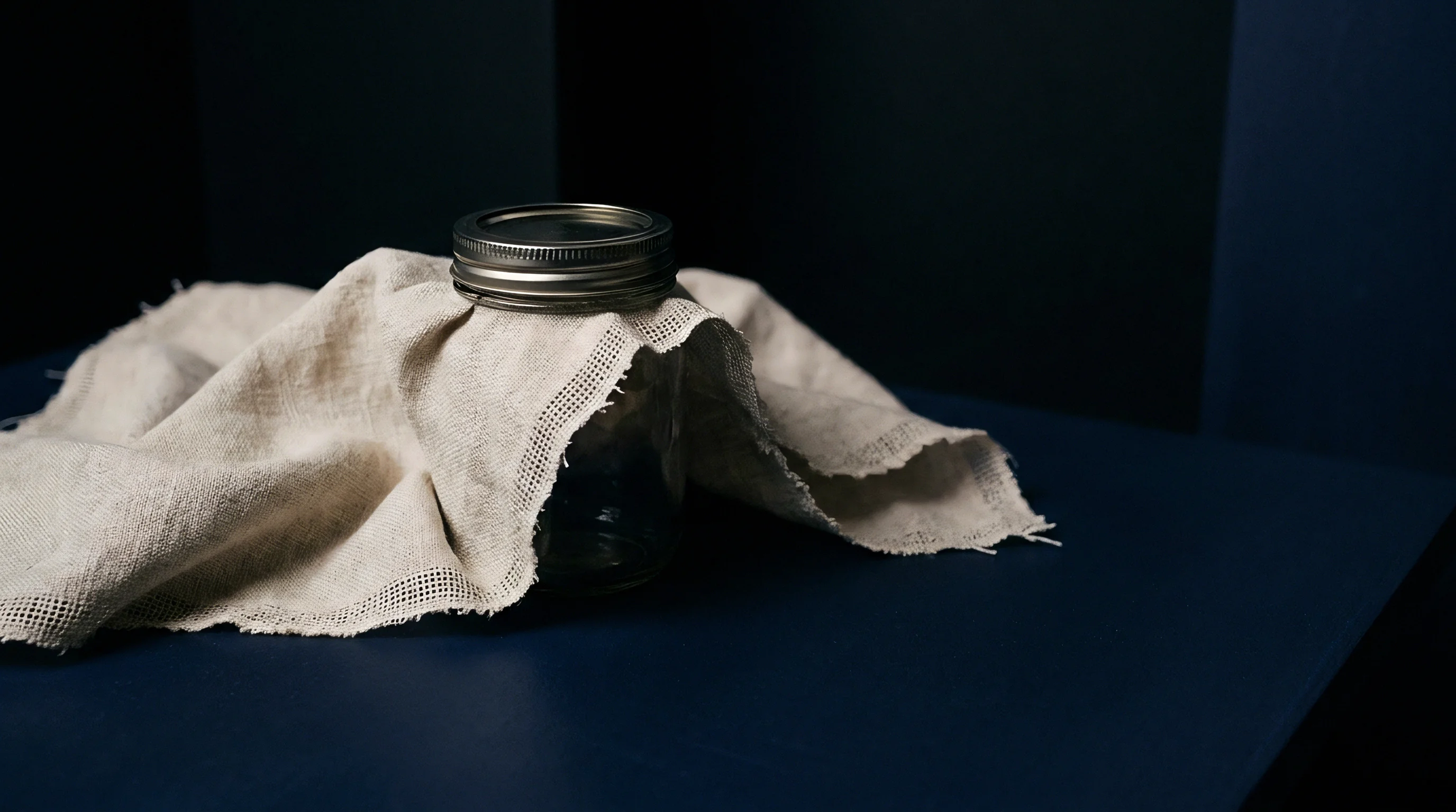 Unbleached woven linen fabric drapes over a clear glass jar resting on a dark blue surface inside a studio.