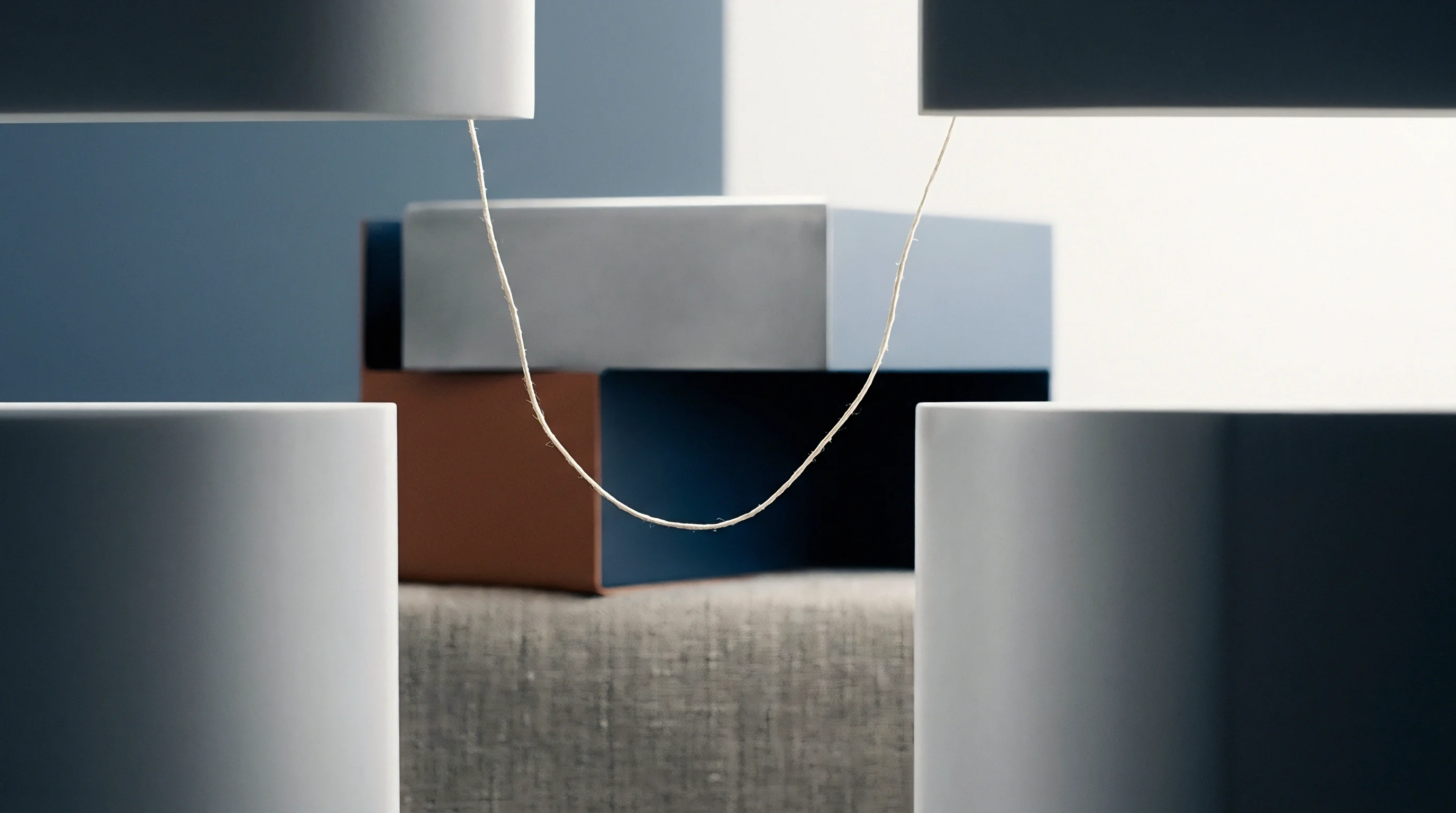 A single natural fibre yarn suspends under tension between geometric blocks above stacked colored containers on a textured textile surface.