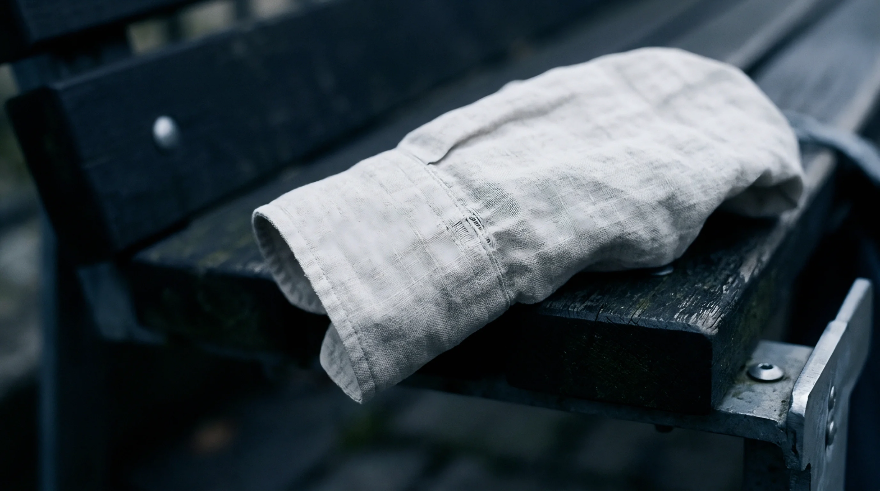 A woven linen sleeve rests discarded across weathered wooden slats of an outdoor park bench under overcast skies.